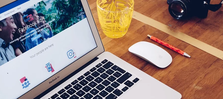 A MacBook Air displaying a Facebook ads page is on a wooden desk. Next to it are a white Apple Magic Mouse, a yellow glass filled with pencils, a red pen, and a camera with a strap.