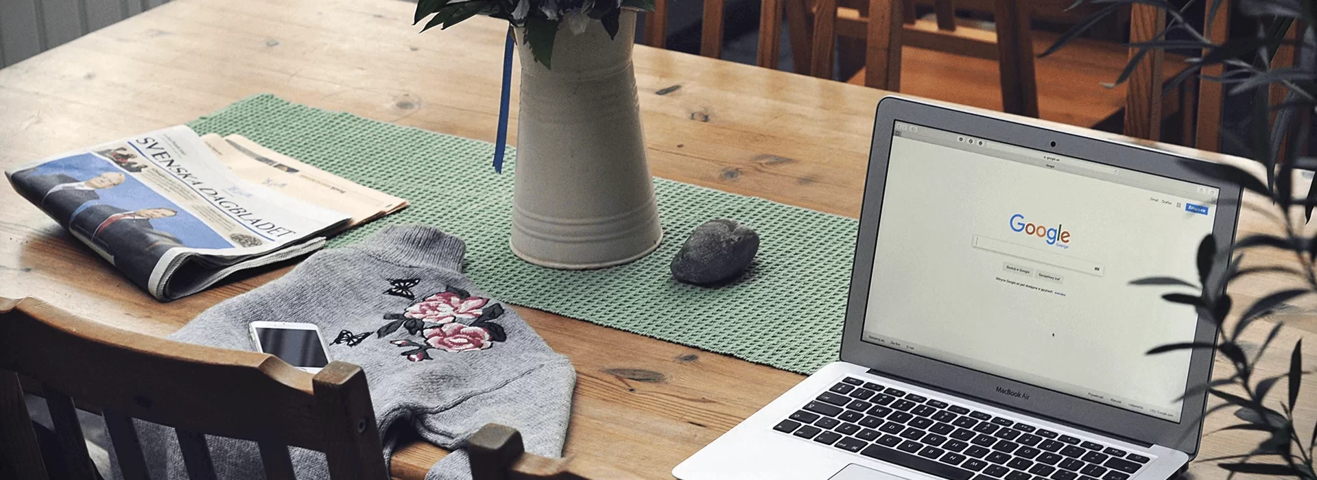 A wooden table setup with a laptop displaying the Google search page, a folded newspaper, a gray floral-embroidered sweatshirt, a smartphone, and a vase with flowers. A green mat is placed under the items, and wooden chairs surround the table.