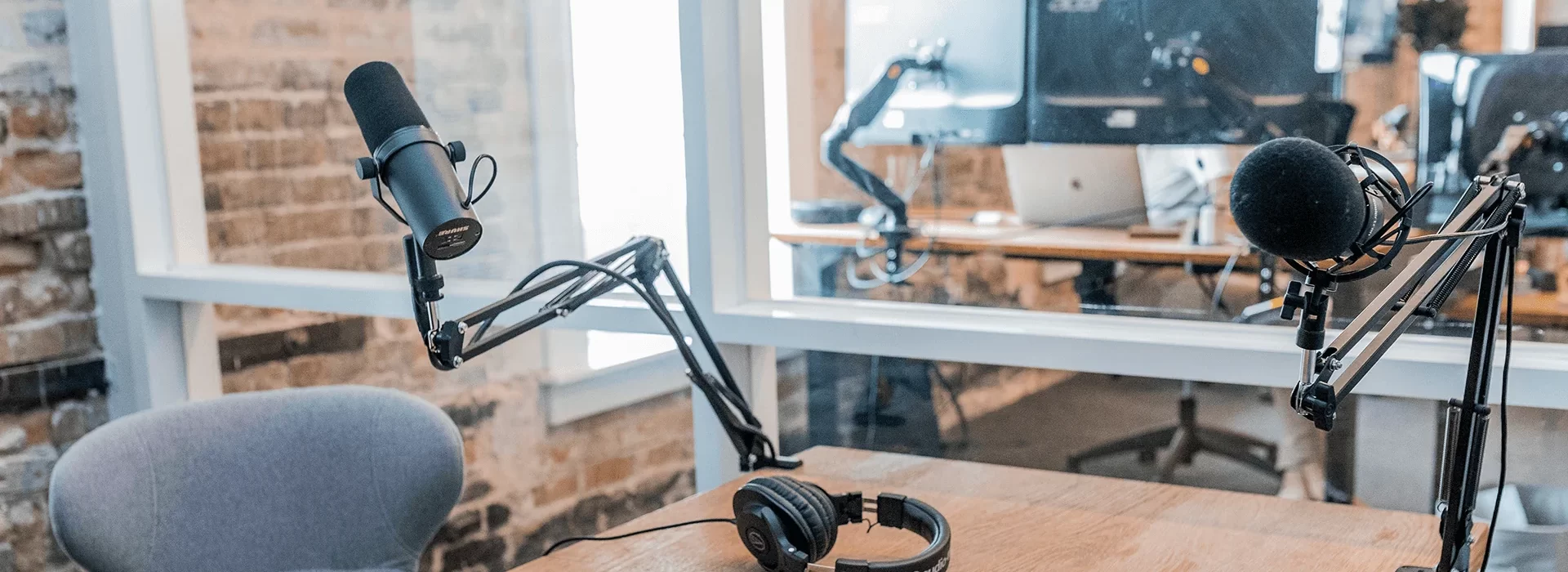 A modern podcast studio setup with two microphones on adjustable arms, a pair of headphones resting on a wooden table, and computer equipment visible in the background through a glass partition. The environment has a professional yet casual feel.