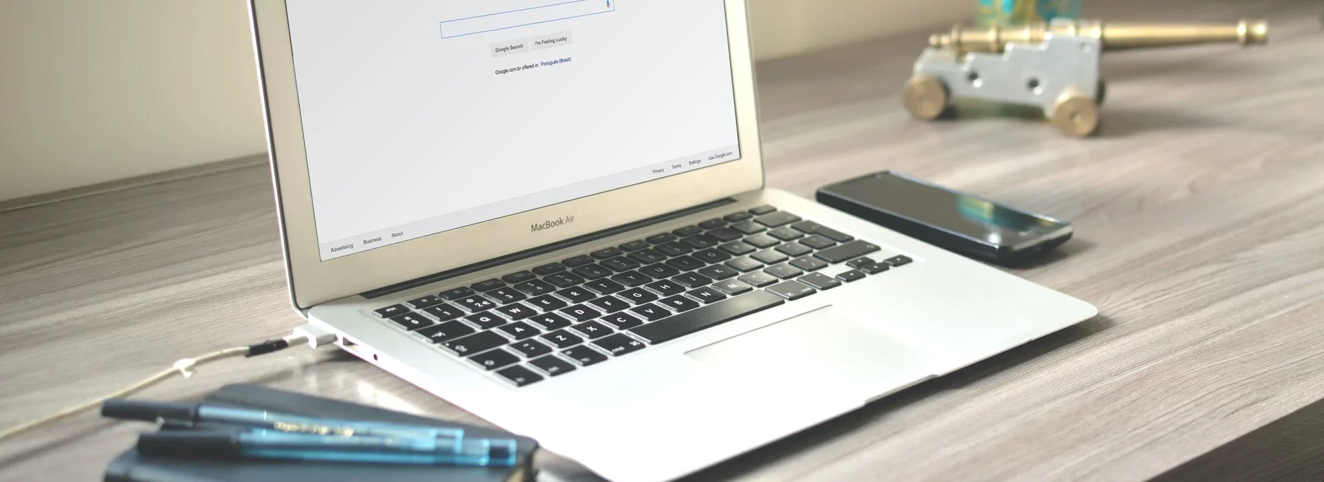A silver MacBook with a blank search engine page open on the screen sits on a wooden desk. The desk also holds a black smartphone, a few blue pens, and a decorative miniature cannon in the background. The scene is well-lit by natural light.