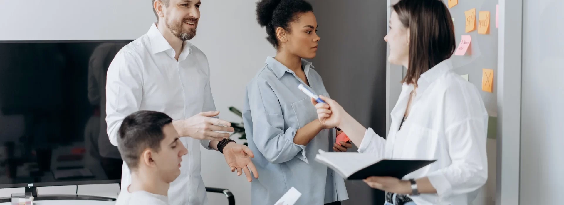 A group of four people are collaborating in an office setting. One person is seated, while the other three engage in discussion, holding notebooks and pens. Sticky notes are visible on a board, suggesting a brainstorming session. All are dressed in casual business attire.
