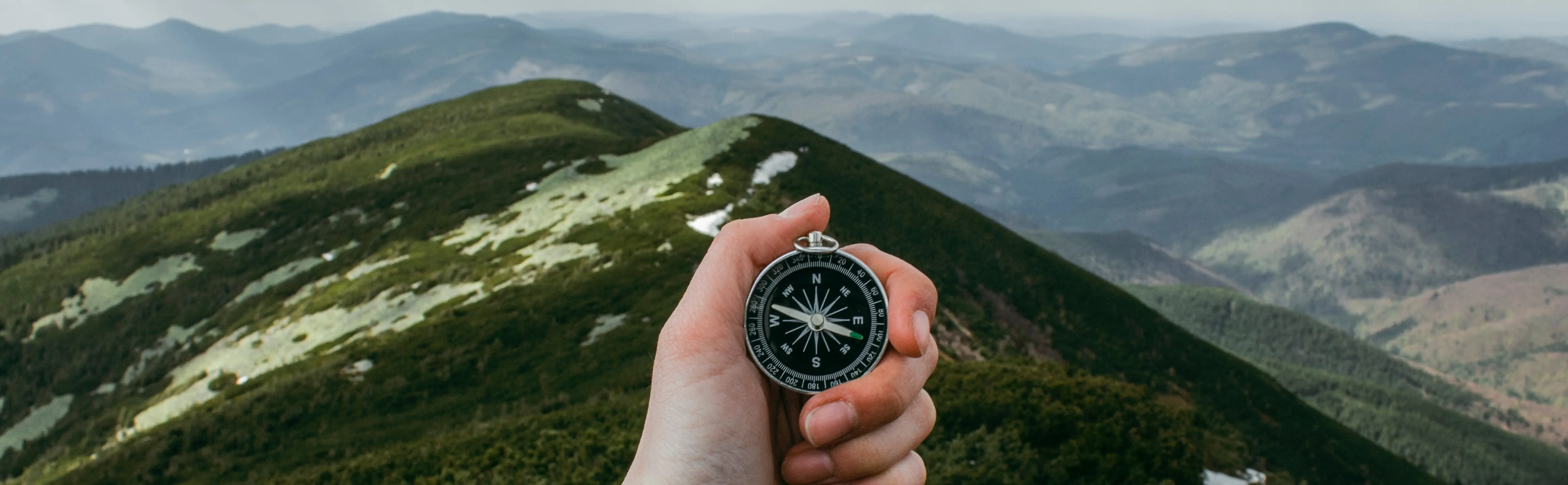 A hand holding a compass, with a view of green, rolling mountains in the background.