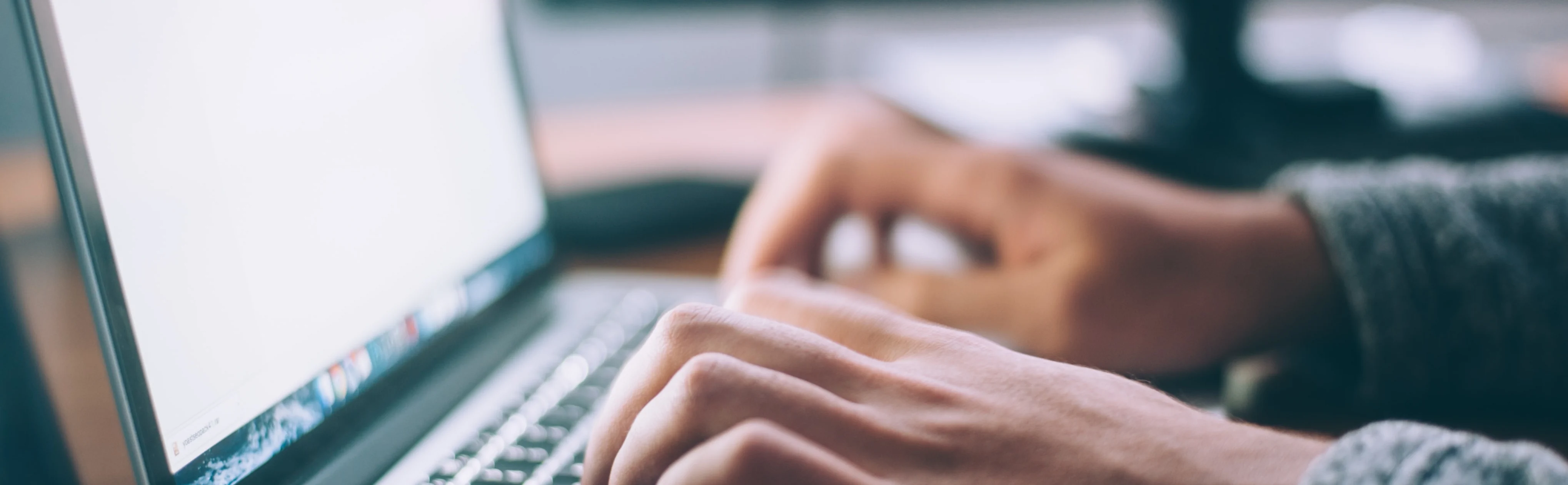 Close-up of hands typing on a laptop keyboard. The screen displays a bright, out-of-focus background.