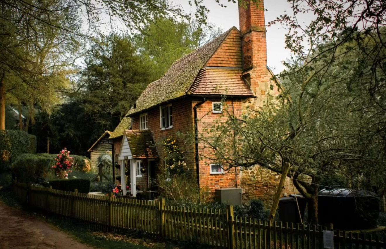 A quaint, two-story brick cottage with a thatched roof, surrounded by trees and a wooden picket fence, in a rural setting.
