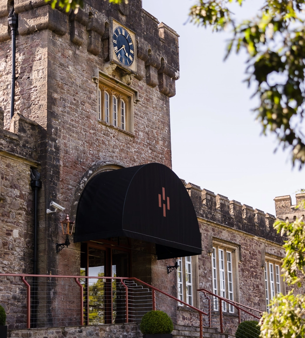 A stone building with a arched entrance canopy, clock, and crenellated battlement. Surrounding foliage is partially visible.
