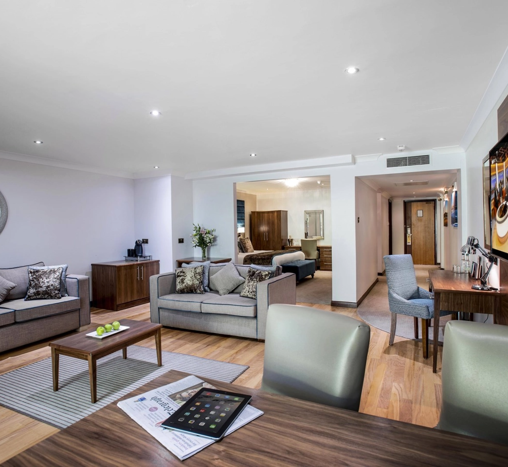 A modern hotel suite featuring a living area with sofas and a coffee table, an adjacent bedroom with a bed, a desk with a tablet, and a dining area with a table and chairs.