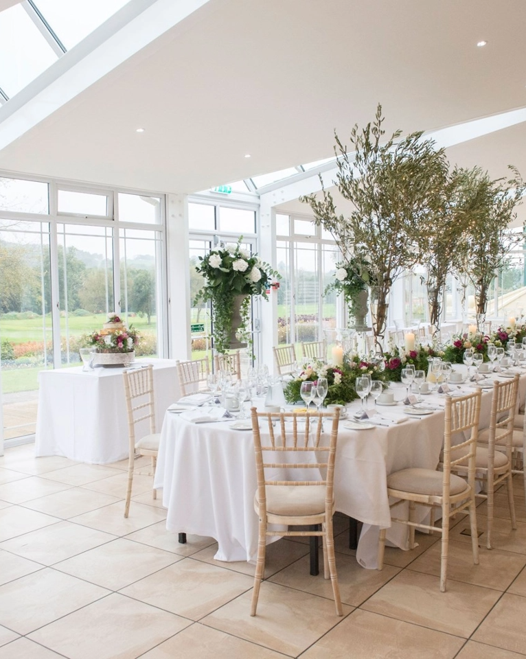 A dining area set up for an event features round tables with white tablecloths, elegant place settings, wooden chairs, floral centerpieces, and a large wedding cake on display in a bright, glass-enclosed room.