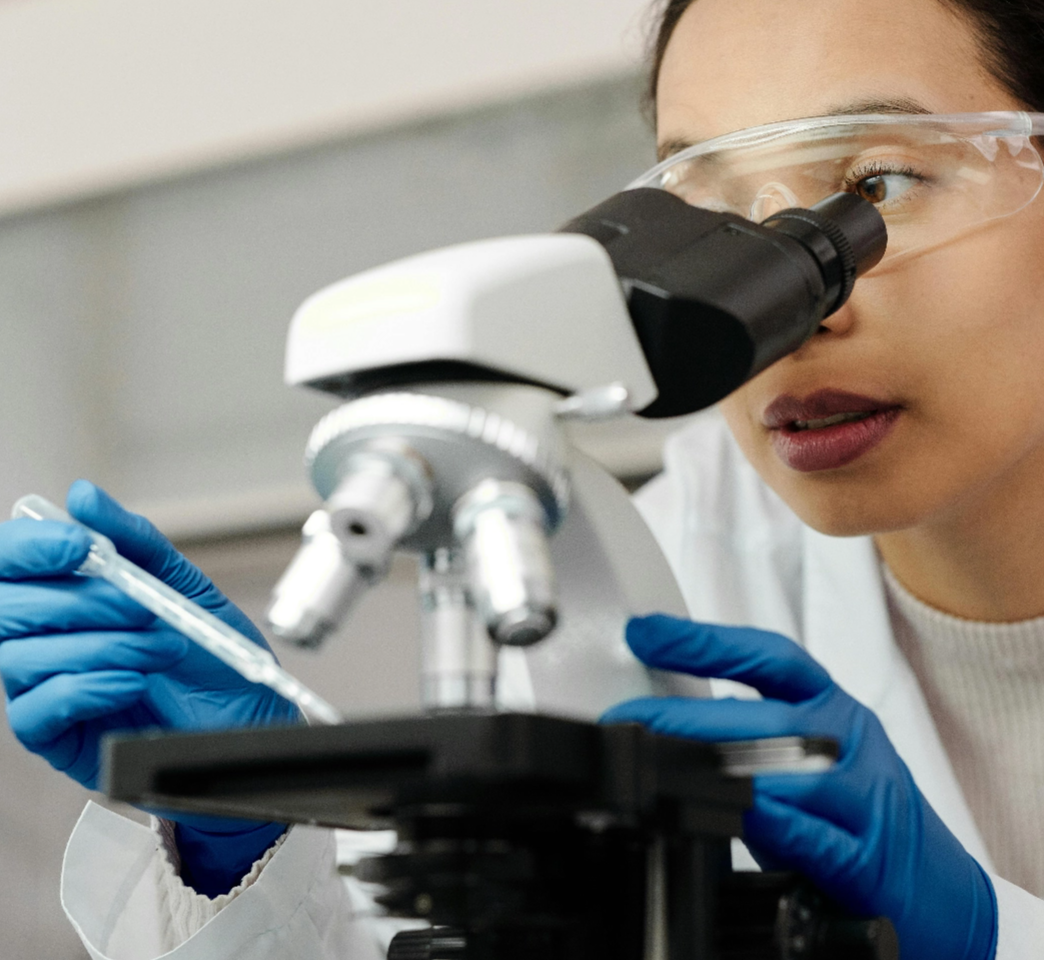 A scientist in a lab coat and safety goggles is using a microscope while holding a pipette.