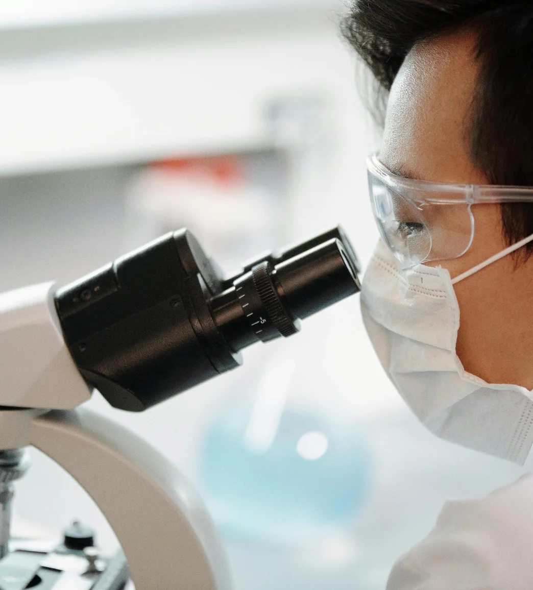 A person wearing safety goggles and a face mask looks into a microscope in a laboratory setting.