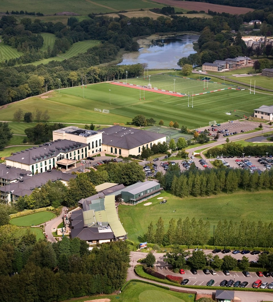 Aerial view of a university campus with sports fields, buildings, parking lots, and surrounding greenery.