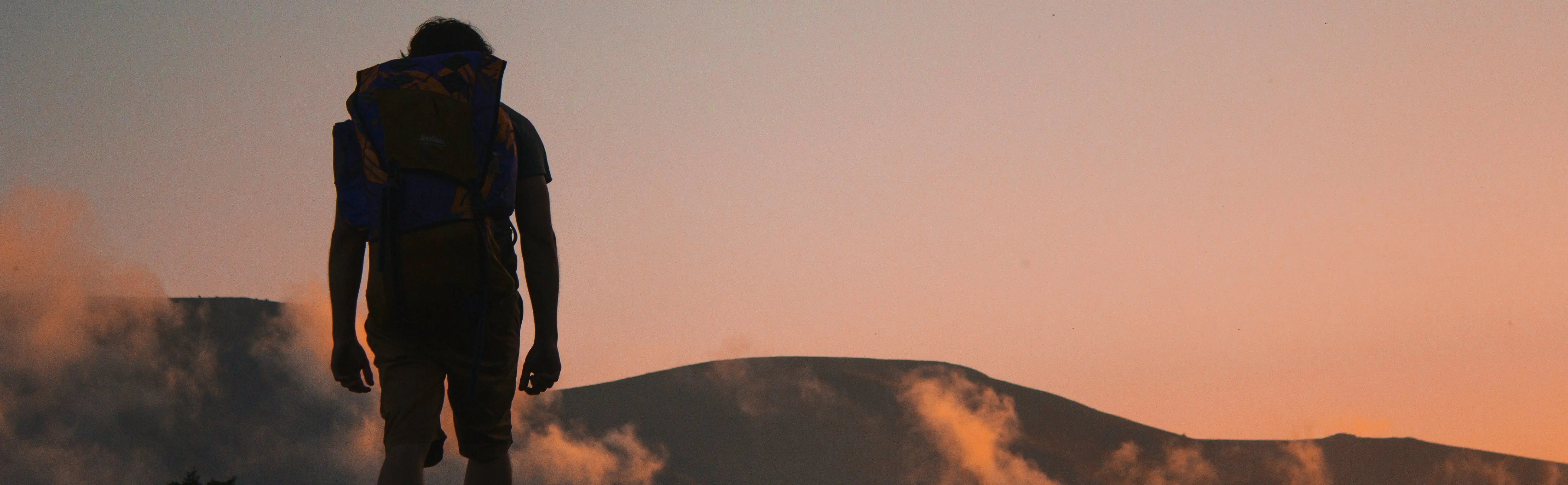 A person with a backpack standing on a hilltop at sunset, with clouds and distant mountains in the background.