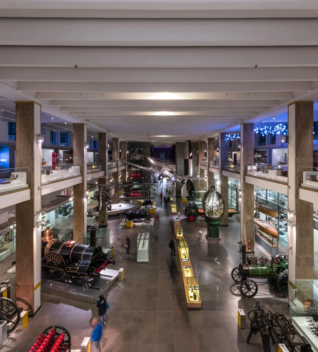 Wide-angle view of an expansive museum exhibit hall featuring various historical machinery and vehicles on display, with visitors walking and observing the exhibits.