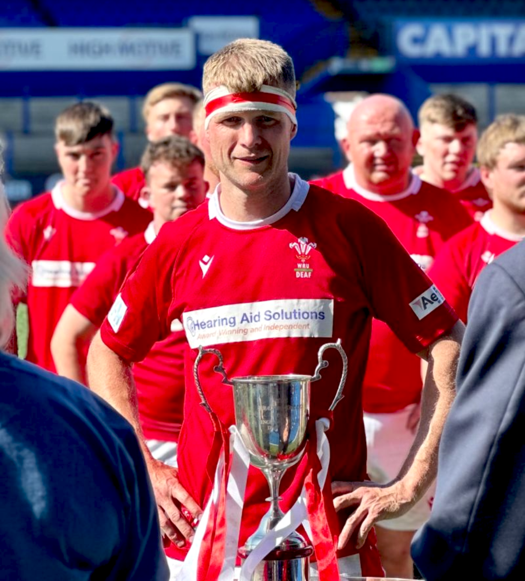 A rugby player wearing a red uniform and a headband stands in front of a trophy with teammates in the background.