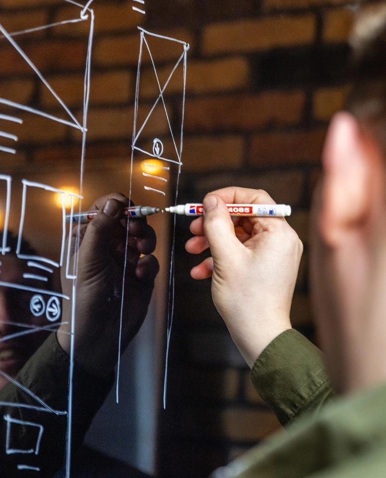 A person draws a website wireframe on a glass board using a white marker, with their reflection visible in the glass.