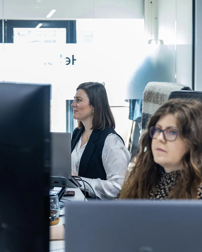 Two women are working at their desks in an office, focused on their computer screens. The background includes frosted glass and a window.