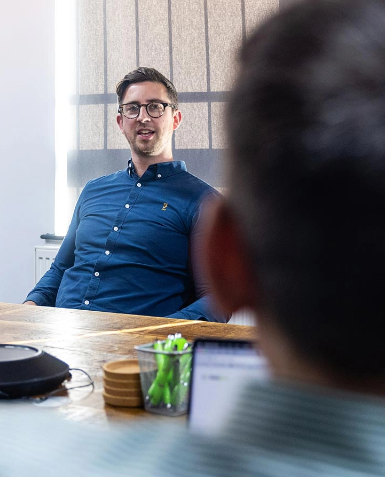 A man in glasses and a blue shirt speaks while sitting at a table with another person in the foreground. The table has various office items, including a laptop and a container of green highlighters.