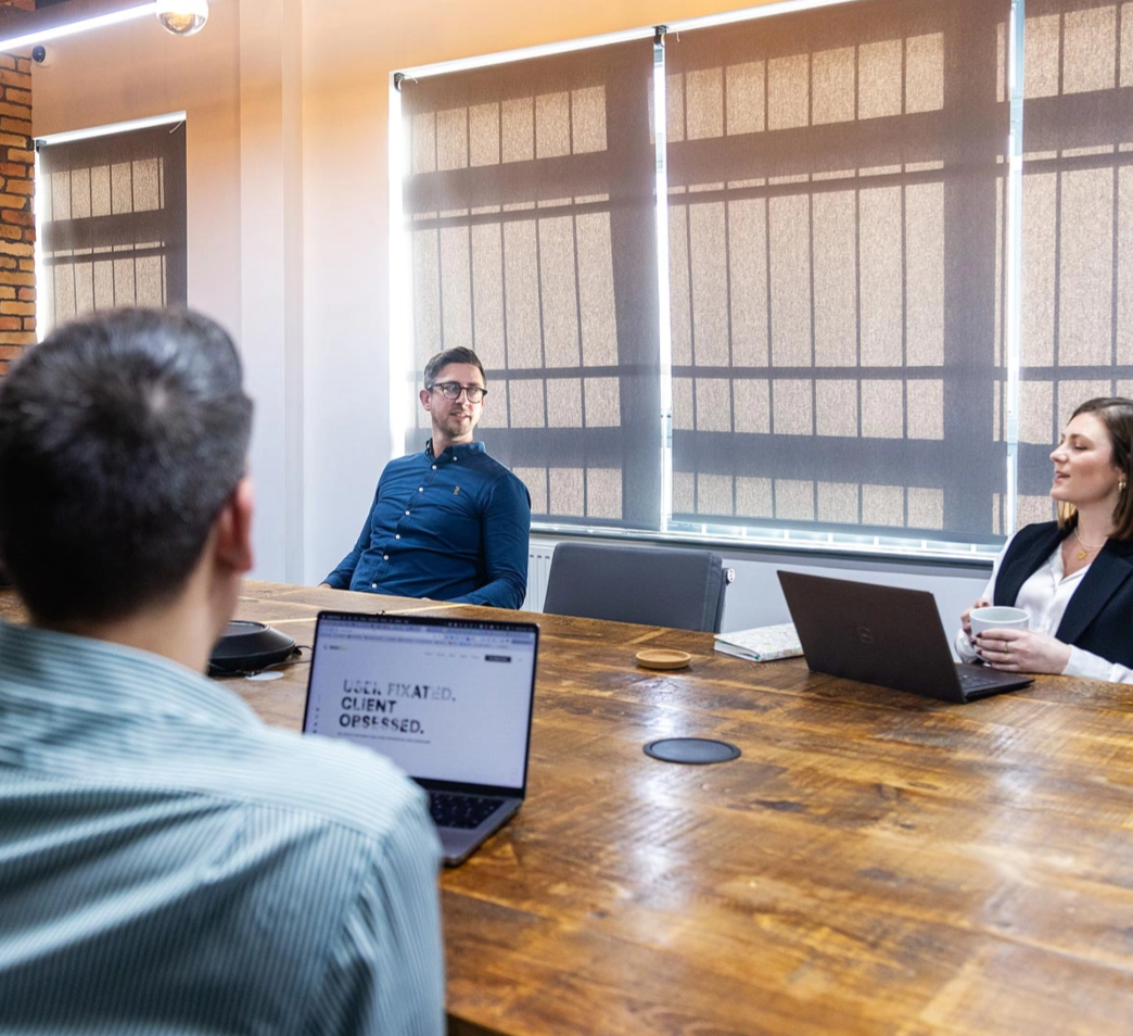 Three people having a meeting in a conference room with laptops on the table. One person is drinking from a mug and others are looking at the speaker.