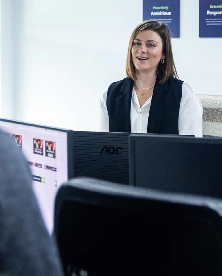 A woman in business attire is sitting at a desk with computer monitors. She is smiling, and there are motivational posters on the wall behind her.
