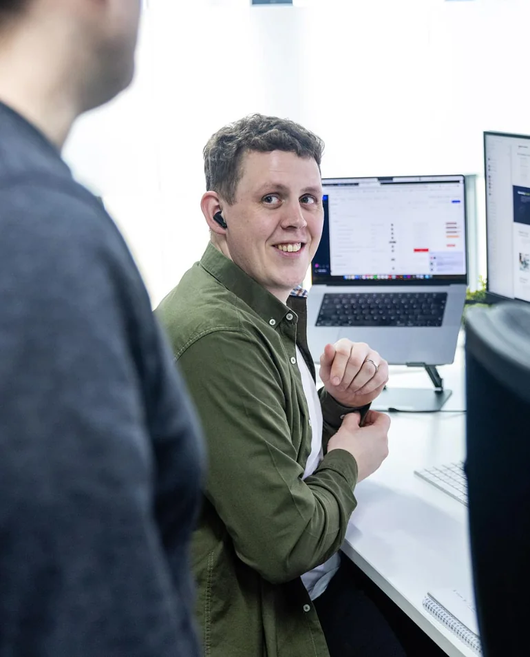 Person with short hair and earbud, seated at a desk with computer screens, looking over their shoulder at another individual standing nearby.