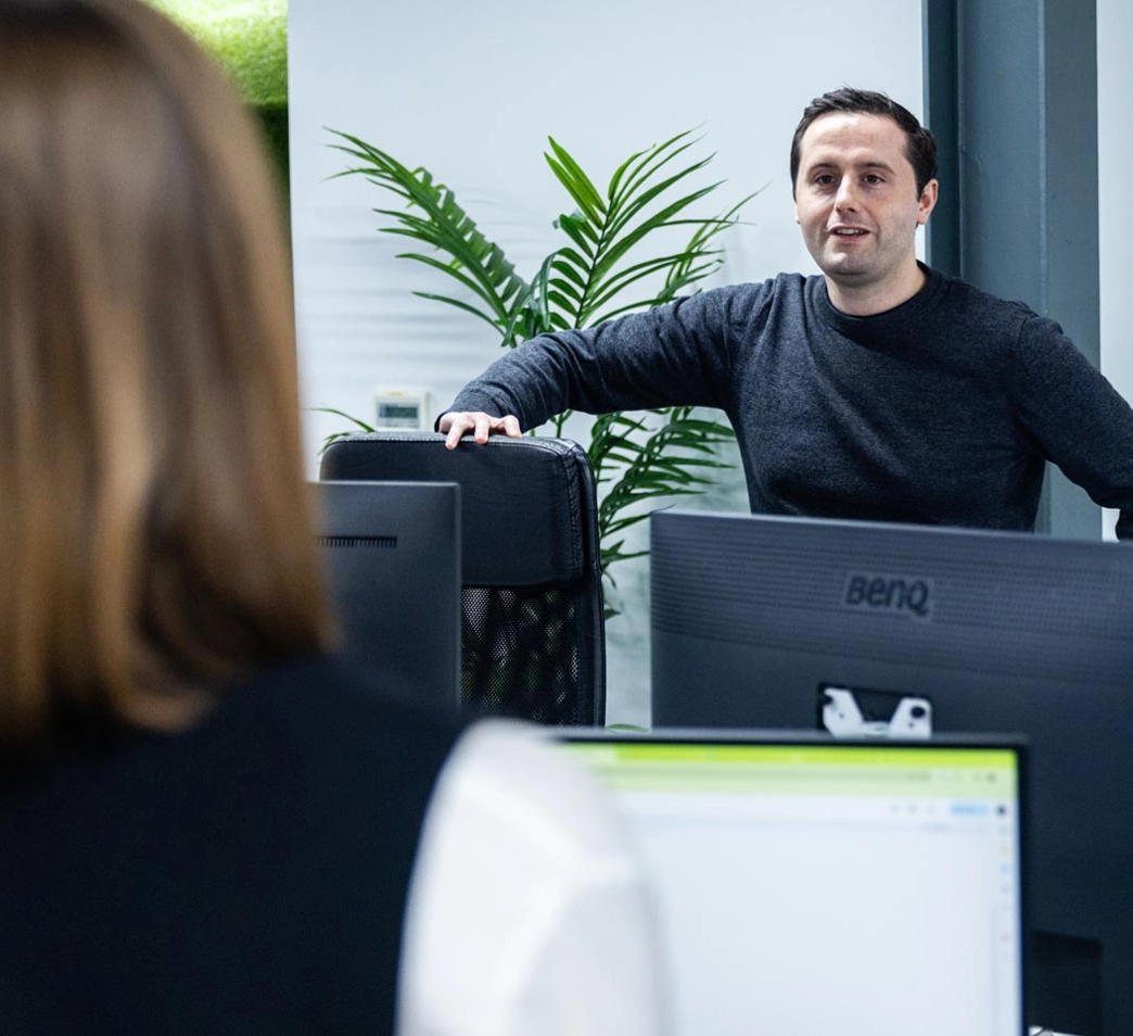 A man in a dark shirt leans on an office chair, talking to a woman with long hair in an office with computers and a plant in the background.