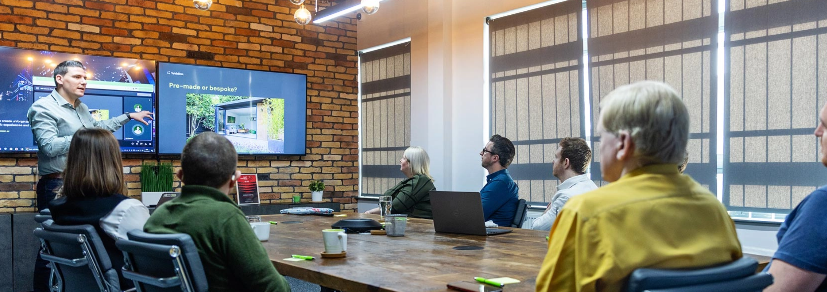 A group of people sit around a conference table in an office, attentively listening to a presentation. The presenter points to a large screen displaying slides.