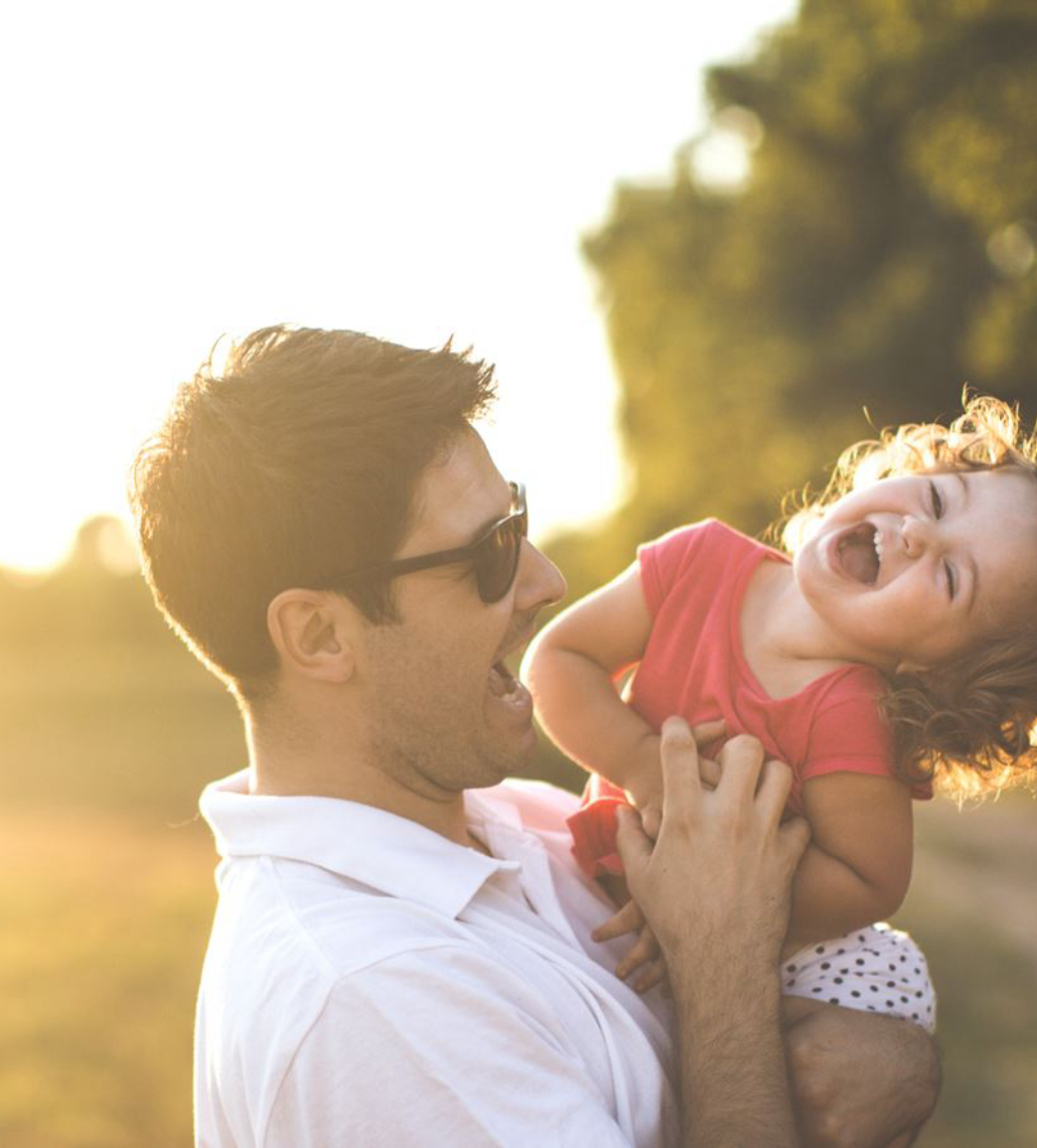 An adult wearing sunglasses holds a laughing child in a red shirt and polka dot shorts outdoors, with sunlight in the background.
