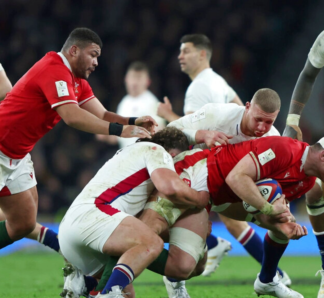 Rugby players from two teams engage in a tackle during a match. One player in a red jersey is being tackled by two players in white jerseys while another player in red observes the action.