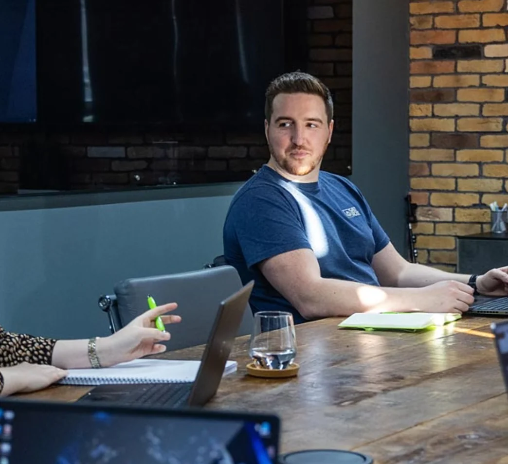 A person in a blue shirt is sitting at a conference table with a laptop, looking to the side. Two other people are partially visible, engaged in discussion. Notebooks and laptops are on the table.