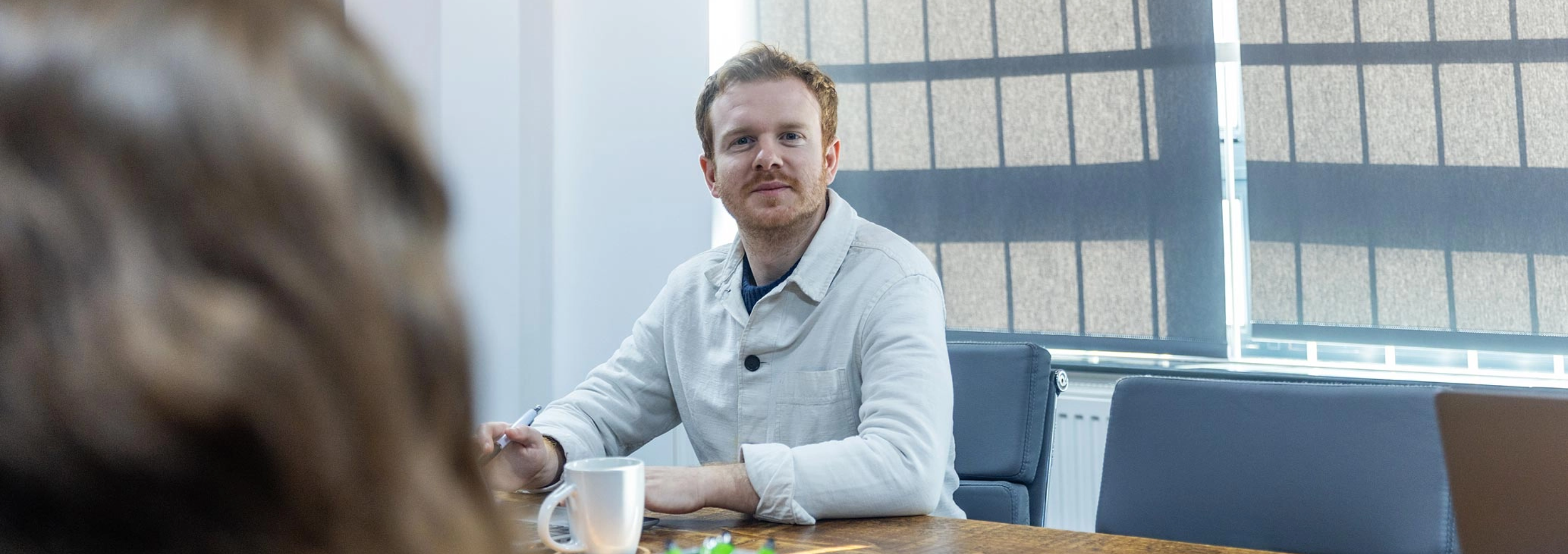 A person with light hair and a beard sits at a conference table with a white mug in front of them, looking forward. Blinds cover the windows in the background.