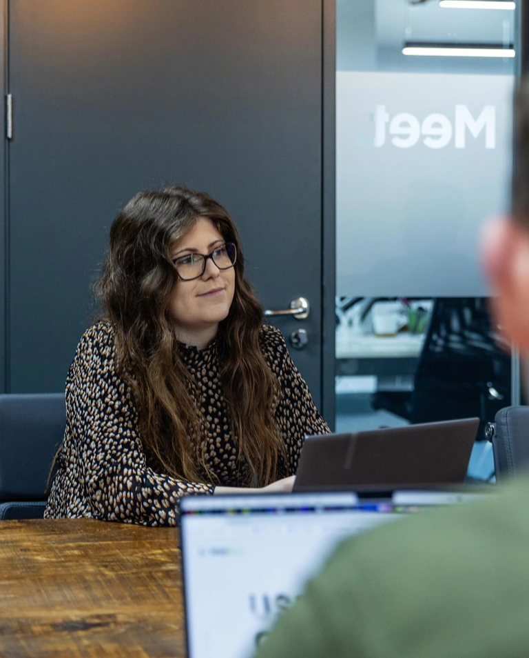 A woman sits at a table with a laptop, looking at a person across from her. The background shows a door and glass wall with partial text "Meet.