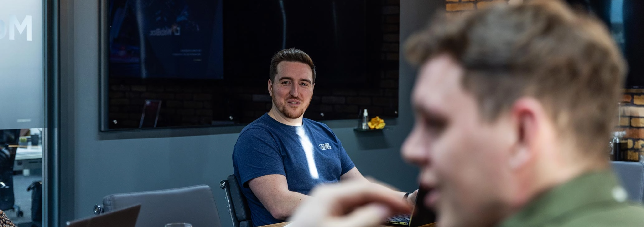 Two men are in conversation in a conference room. One man in a blue shirt is in focus, while the other, in the foreground, is blurred. A presentation screen is visible in the background.