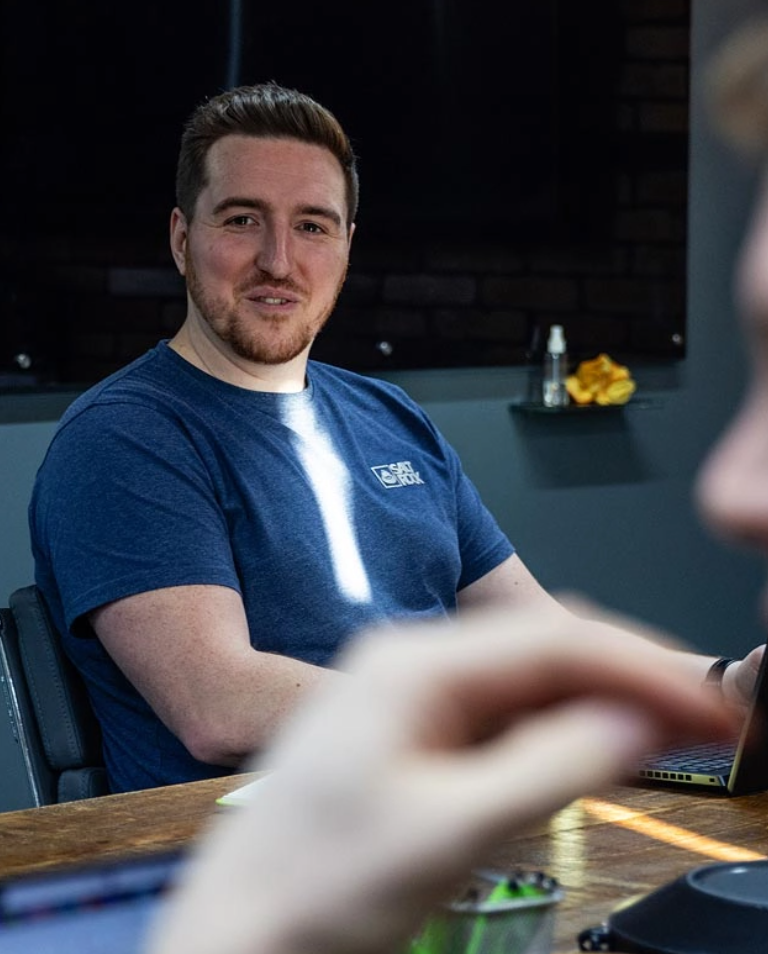 A man with short hair and a beard wearing a blue T-shirt is sitting at a table, looking at the camera. He is indoors with dark walls and a shelf with some items behind him.