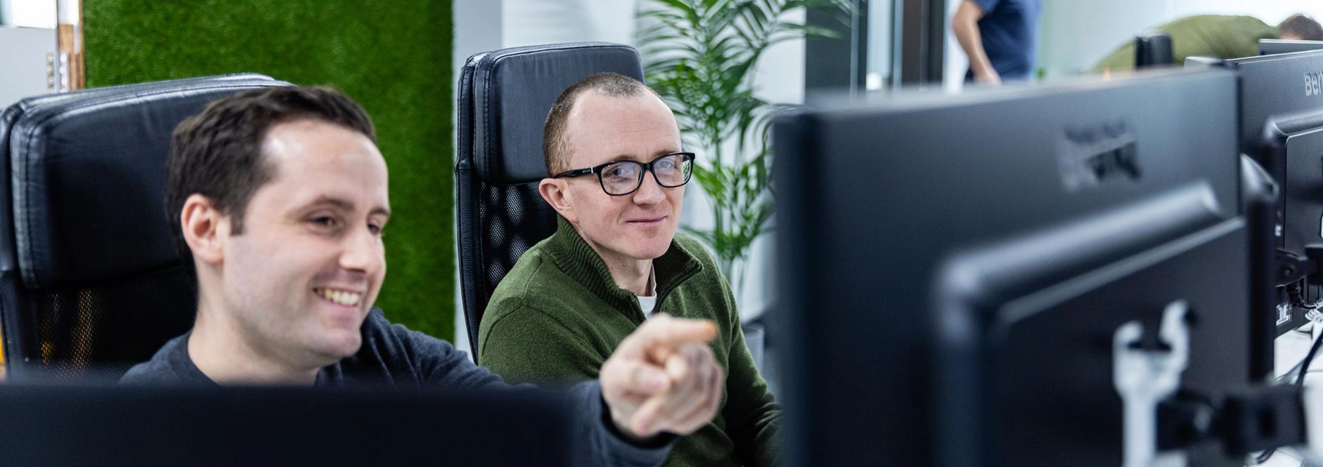 Two men sit in office chairs in front of computer monitors. One man is pointing at something on the screen while the other is watching attentively.