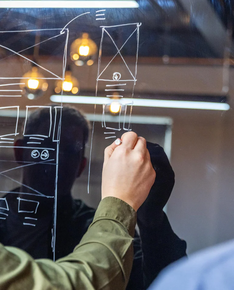 Person drawing architectural designs on a transparent board with a white marker, with blurred lights and modern interior in the background.