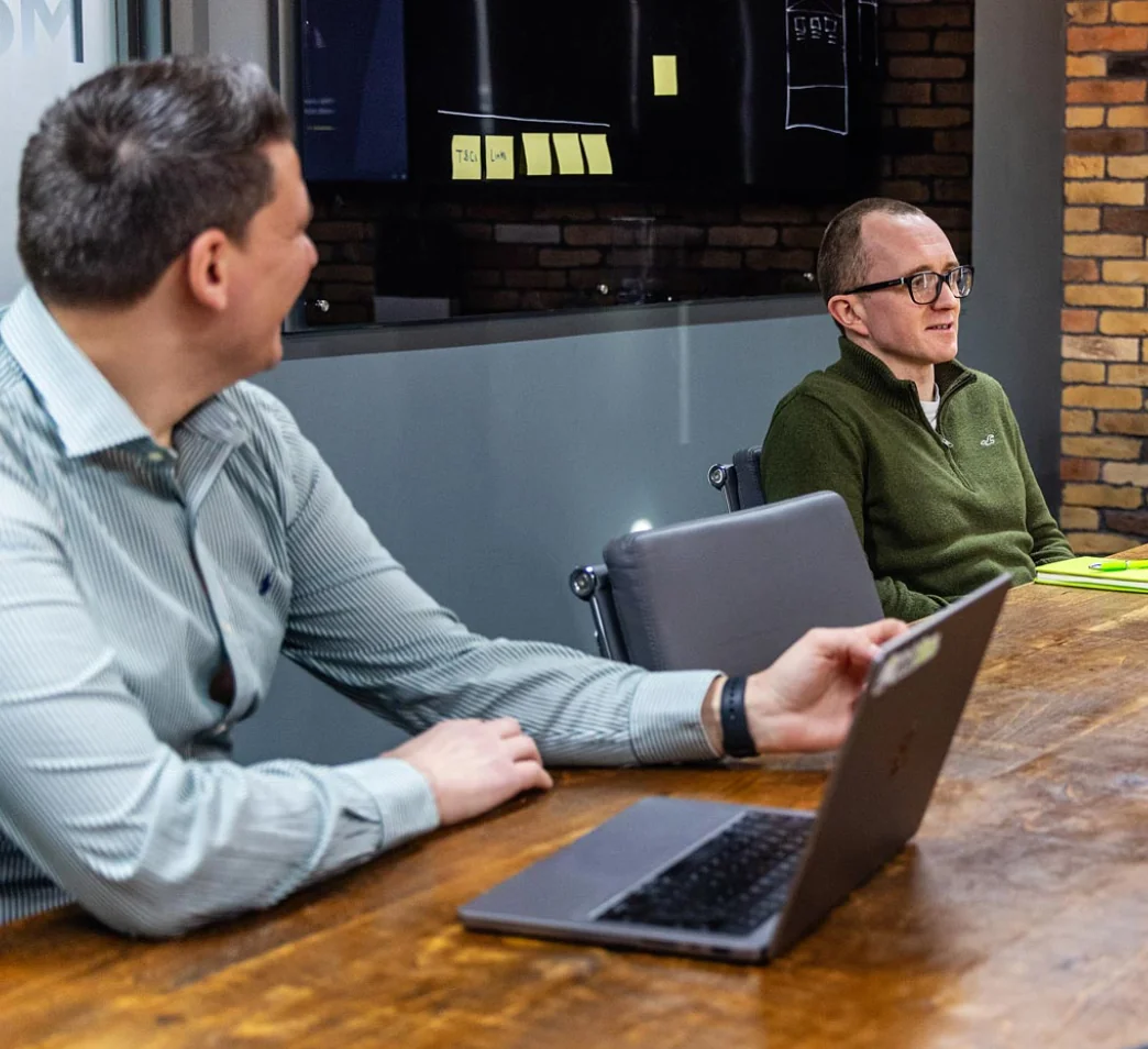 Two men sitting at a wooden conference table, one is talking while the other listens. A laptop and sticky notes are on the table. Brick wall and large screen in the background.