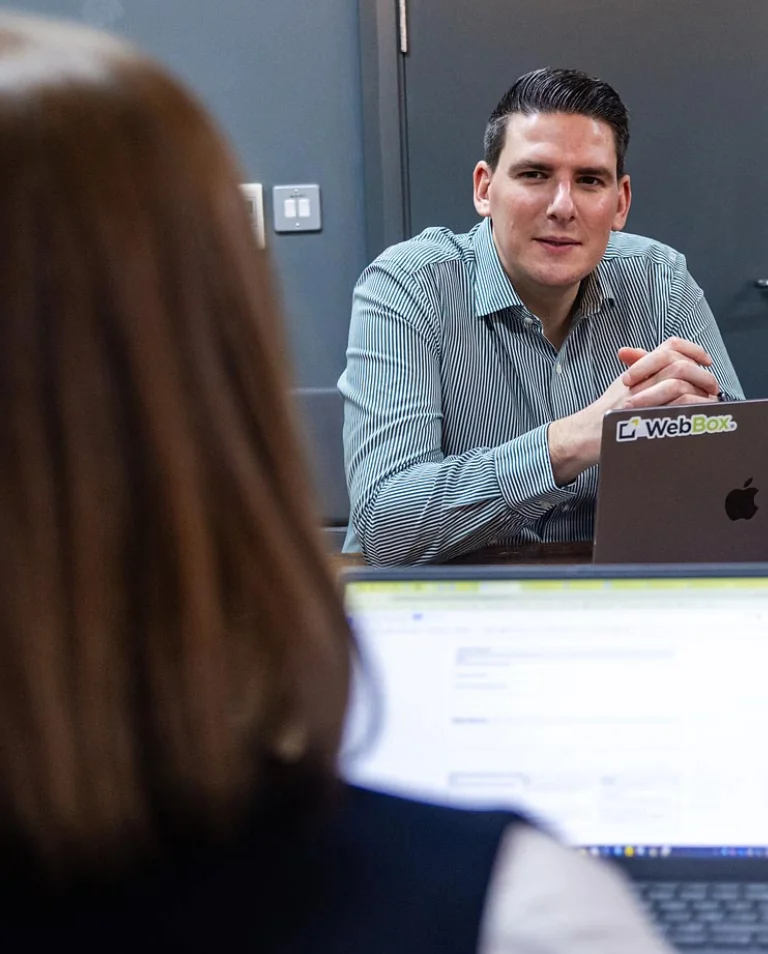 Two people are engaged in conversation. One person is facing the camera, sitting behind a laptop with a "WebBox" sticker. The other is seated across, seen from behind, with a laptop screen visible.