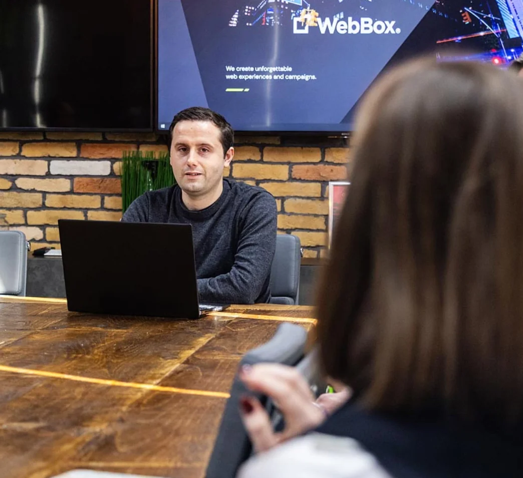 A man sits at a table with a laptop in a conference room, speaking to a woman. A screen behind him displays a "WebBox" logo and text.