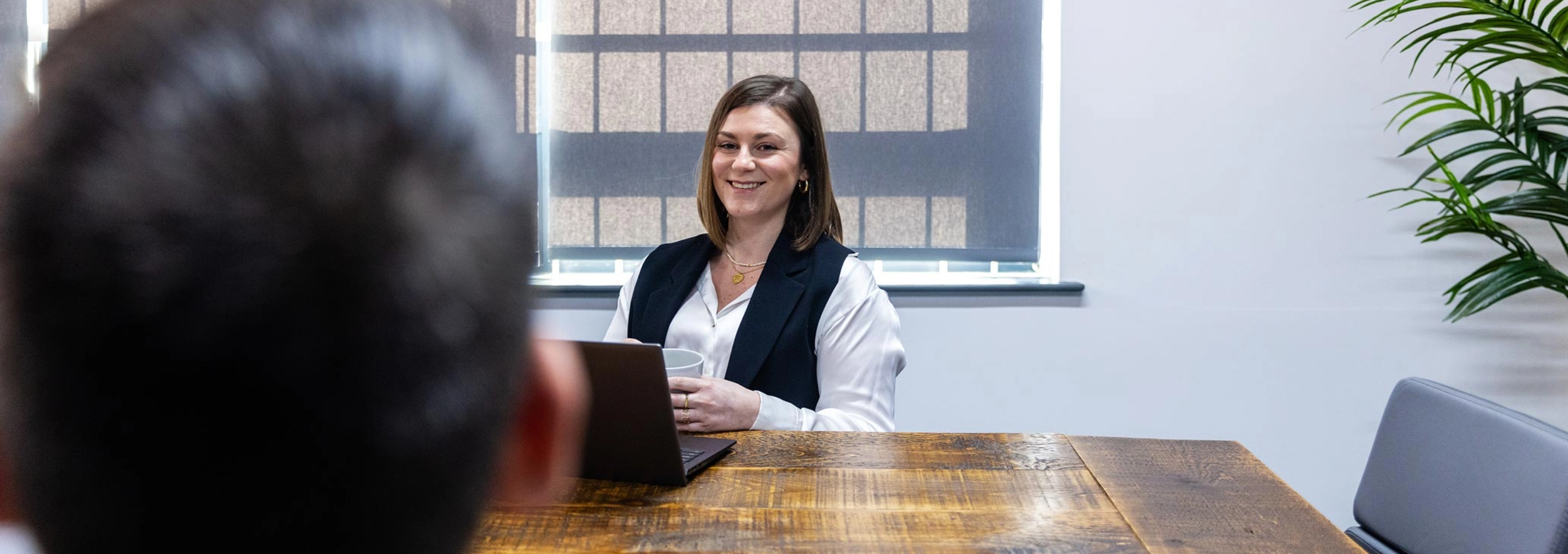A woman sits at a wooden table with a laptop, smiling and talking to a person whose back is to the camera.