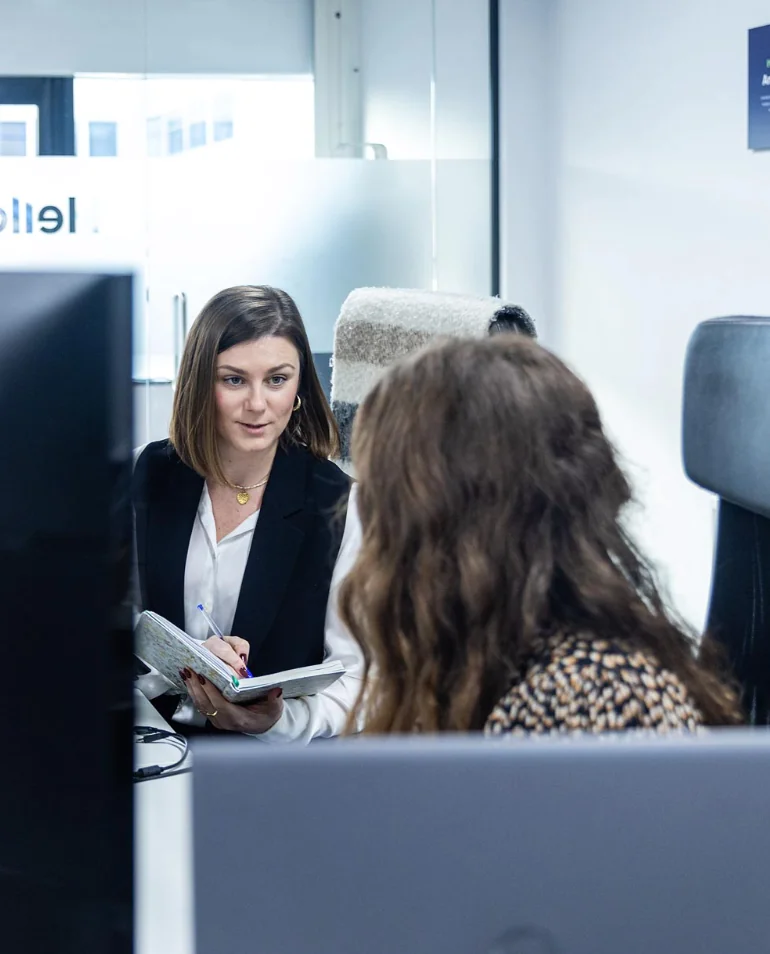 Two women having a conversation at an office desk, one holding a notebook and pen, the other facing away, with computer monitors visible in the foreground.