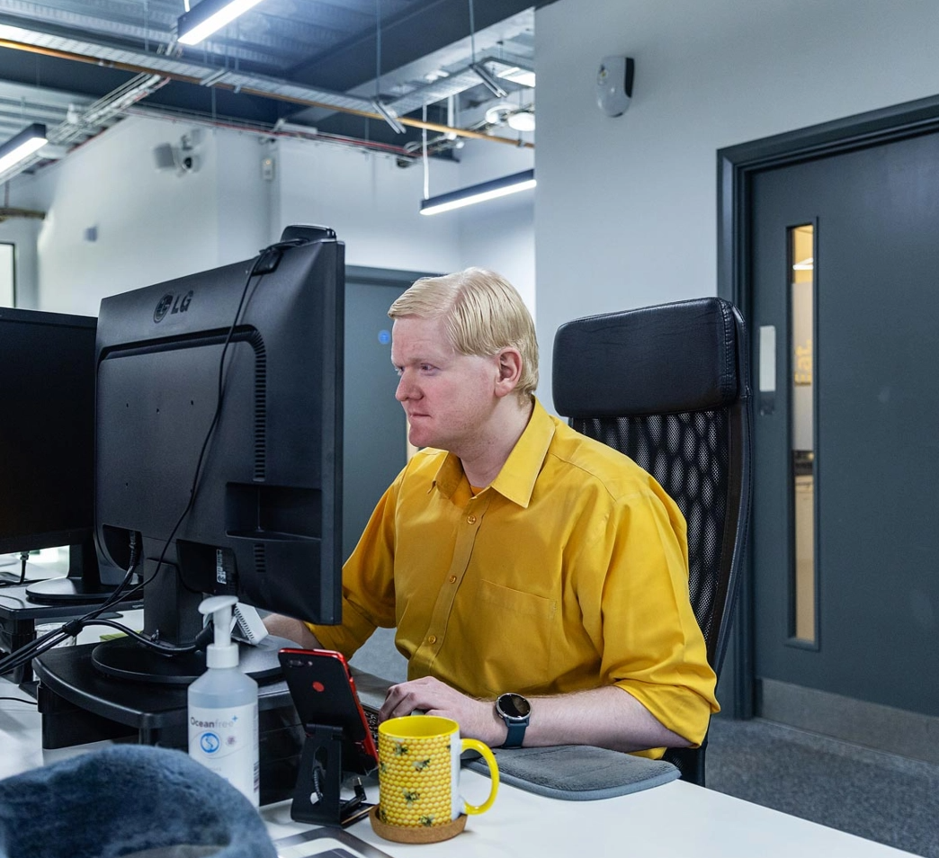 A person wearing a yellow shirt is working at a computer in an office environment. A sanitizer bottle, phone, and a yellow mug are on the desk.