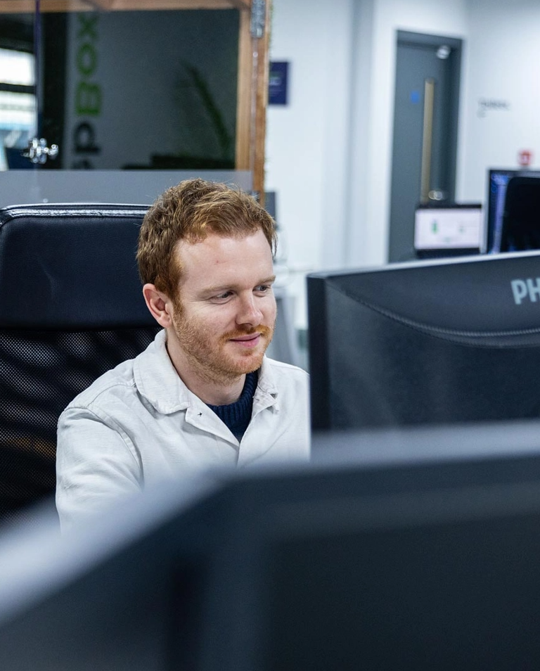 A person with red hair and a beard is sitting at a desk, working on a computer in a modern office environment.